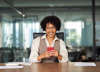 Happy young professional African American business woman executive holding cellphone using mobile looking at camera with cell phone tech in hands sitting at desk in office working, portrait.